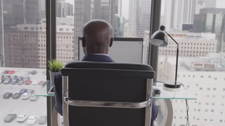 Male sitting at a small desk in front of a window overlooking the city while working on a computer