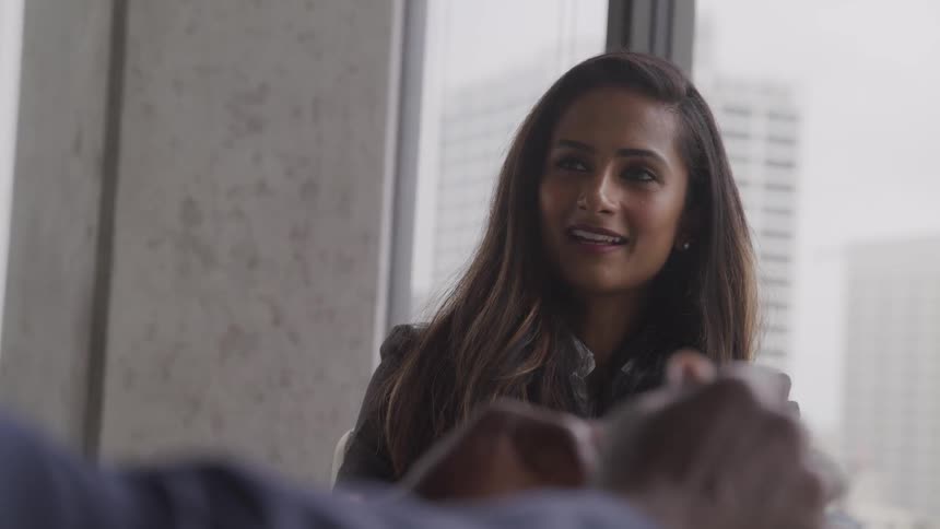 A man and woman sitting in an office talking with a window and buildings in the background
