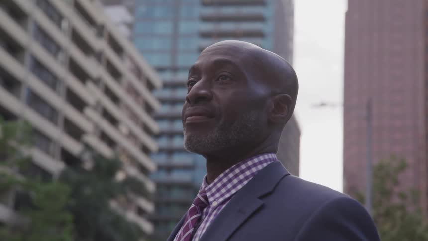 A black man in a suit stands downtown and looks off into the distance