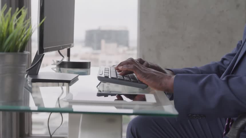 Old black businessman sitting at the glass desk types on the keyboard in the office