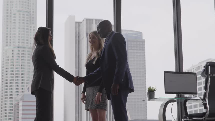 Three business people greeting each other and shaking hands in an office with downtown area seen through the large glass windows