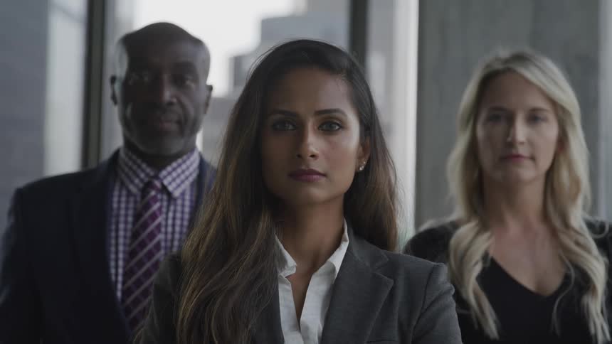 Three people in professional work attire look straight ahead at the camera with windows behind them