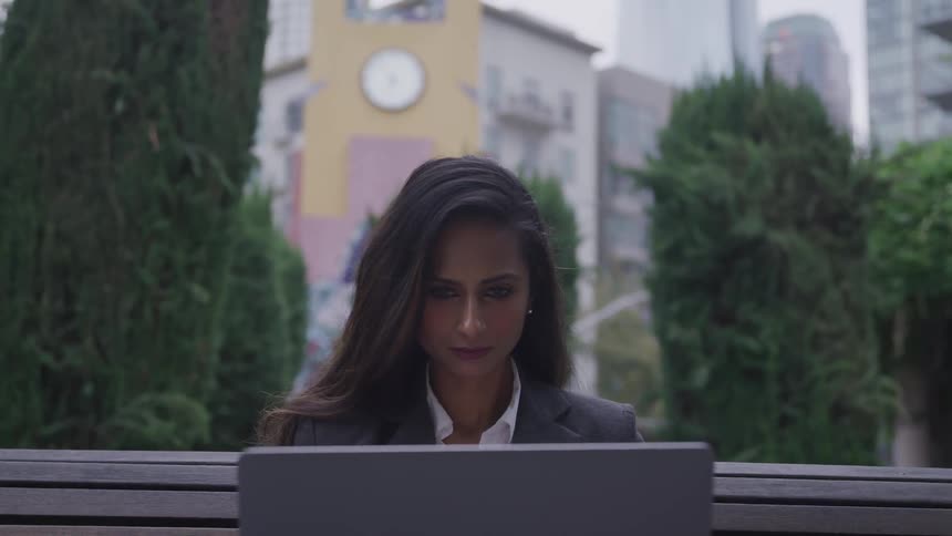 A young woman in business attire sits outdoors on a bench while focusing intensely at her laptop