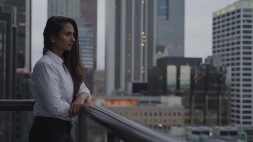 A woman stands with her arm resting on the railing of a balcony overlooking the city