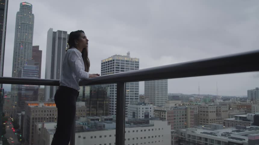 A woman leans against a high rise balcony railing looking towards the city