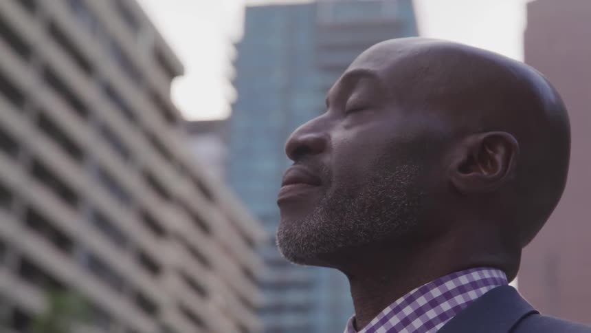 A man stands with his eyes closed and head raised in a city surrounded by buildings