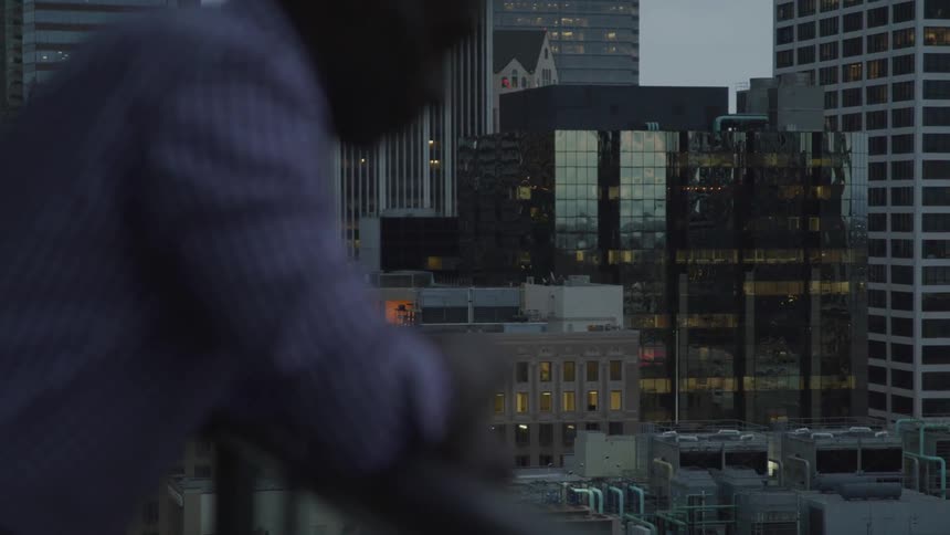 A man stands on a balcony in a large city surrounded by buildings