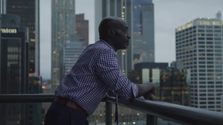 A black man stands on a balcony and looks out at the city