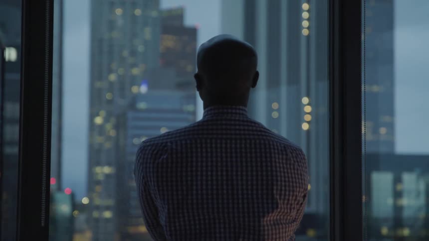 Black businessman looks out the window at the tall buildings in the office at dusk