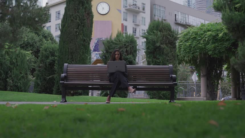 A young woman is sitting on a park bench working on her laptop