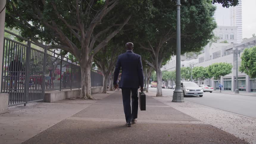 A man carrying a briefcase walks down a city sidewalk as cars pass by