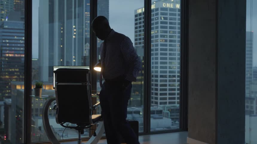 Old black businessman walks in the office with the view of tall buildings at dusk