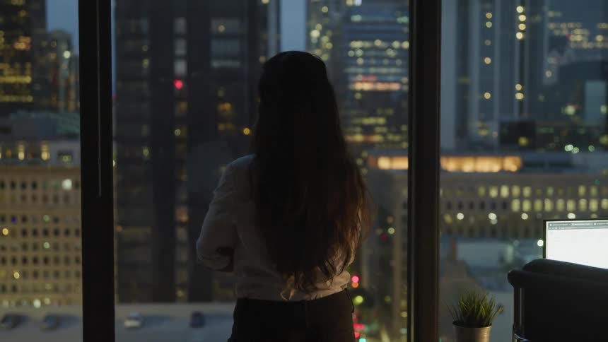 A woman looks out a window at skyscrapers in a city apartment at night