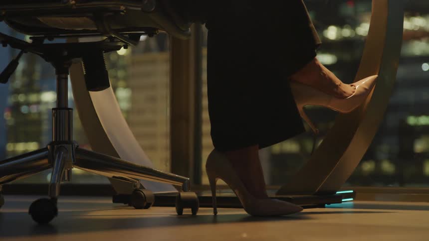 Businesswoman wearing high heels sits at the desk in the office at night