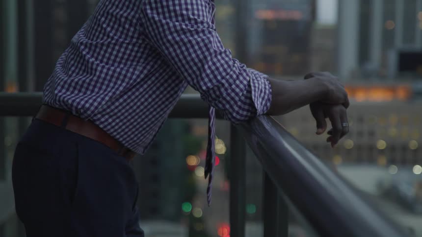 A black man leans on a balcony railing and looks at the city below him