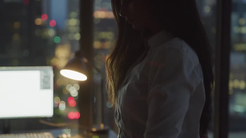 A woman stands by a desk as the city lights shine outside her office window