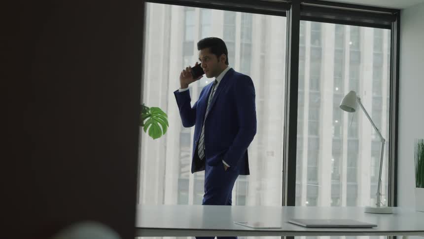 A man in a suit pacing before a window in a skyscraper while speaking on a smartphone
