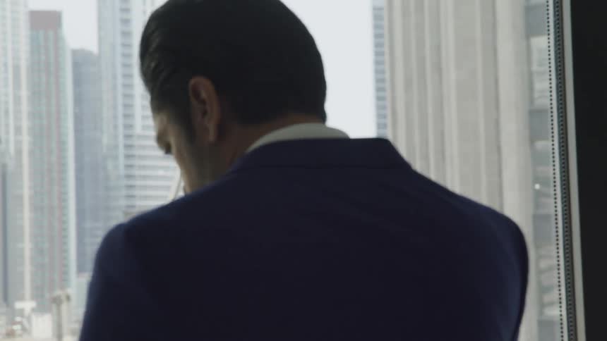 A man in a suit talking on a smartphone while pacing before the window in a skyscraper