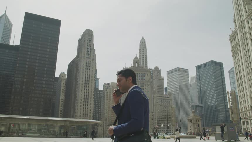 Businessman wearing a blue suit talks to someone on the phone in the city square with the view of tall buildings during the day