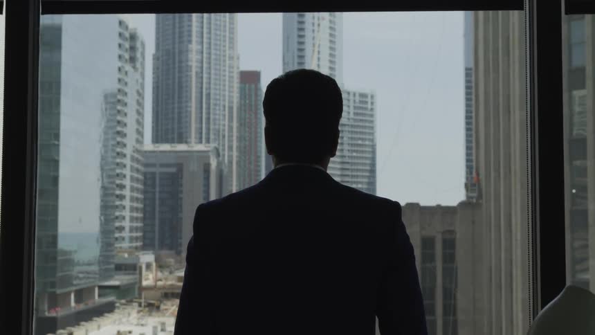 A man stands at a window looking out over an urban area