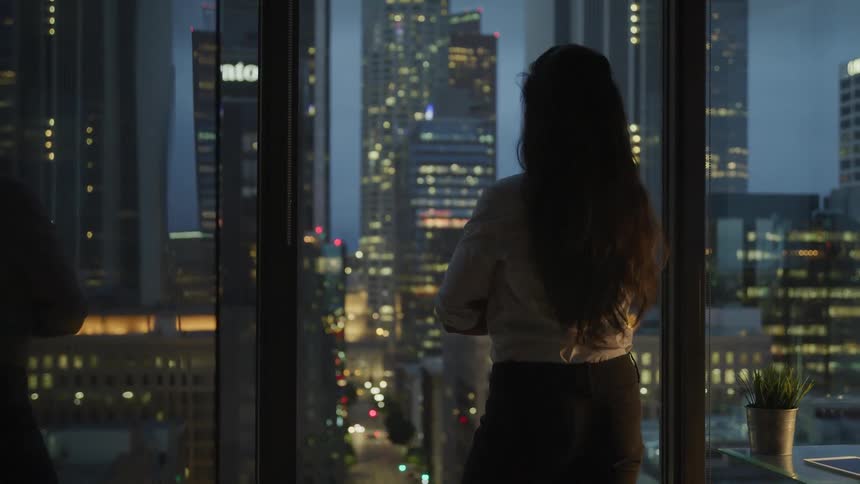 Female arms are crossed in front of her as she looks out her office window at the city lighting up during sunset