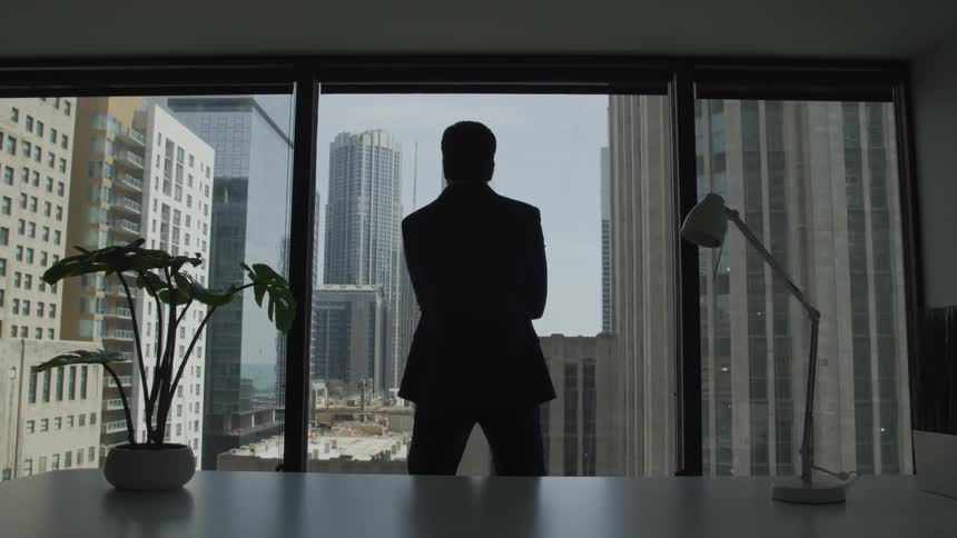 The silhouette of a man in an office facing the windows and looking out at the city