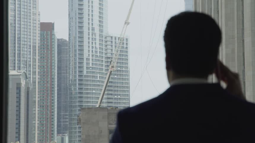 A man standing before a window in a skyscraper listening to a smartphone