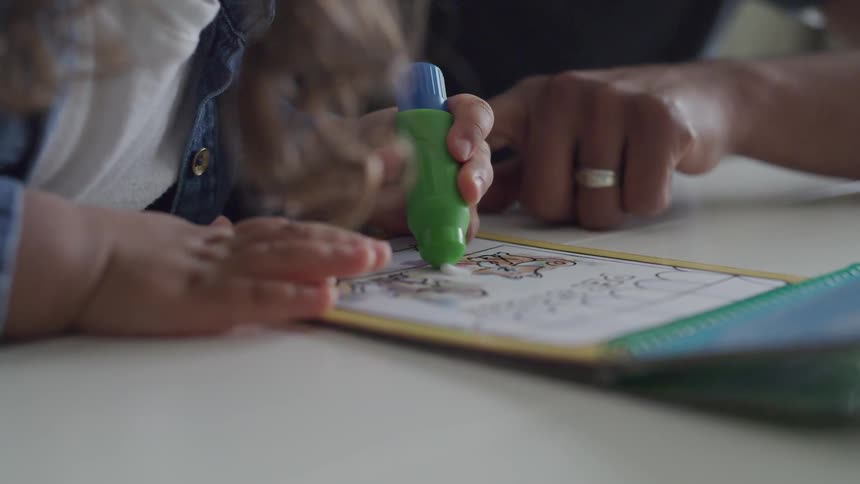 A toddler writes on a whiteboard under close supervision of an adult