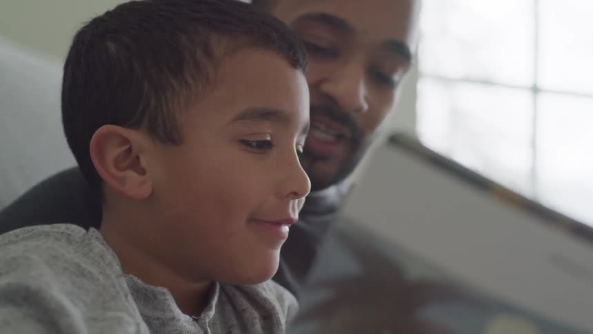 A boy sits with his dad who is reading a book to him