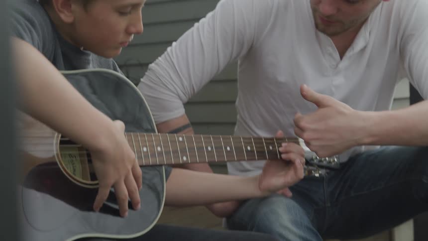 A young boy plays a guitar as a man shows him strings