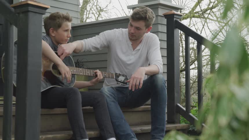 A man teaching a boy how to play the guitar while the two sit on the steps before a house