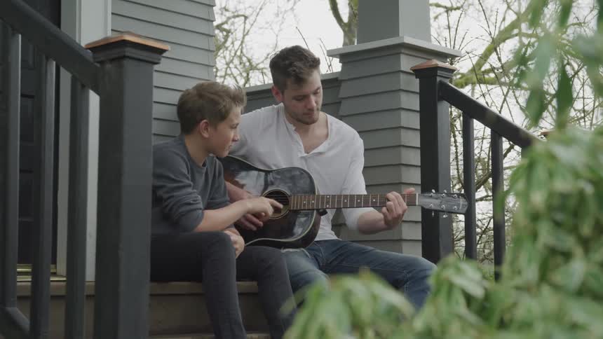 A young boy and his brother sit outside and play the guitar together