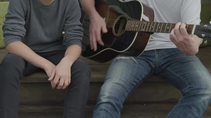 A man plays the guitar sitting next to another man on a step outside