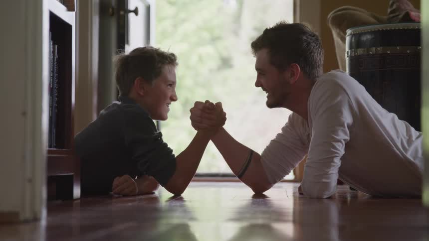 A grown man and a child arm wrestle while lying on the floor of a house