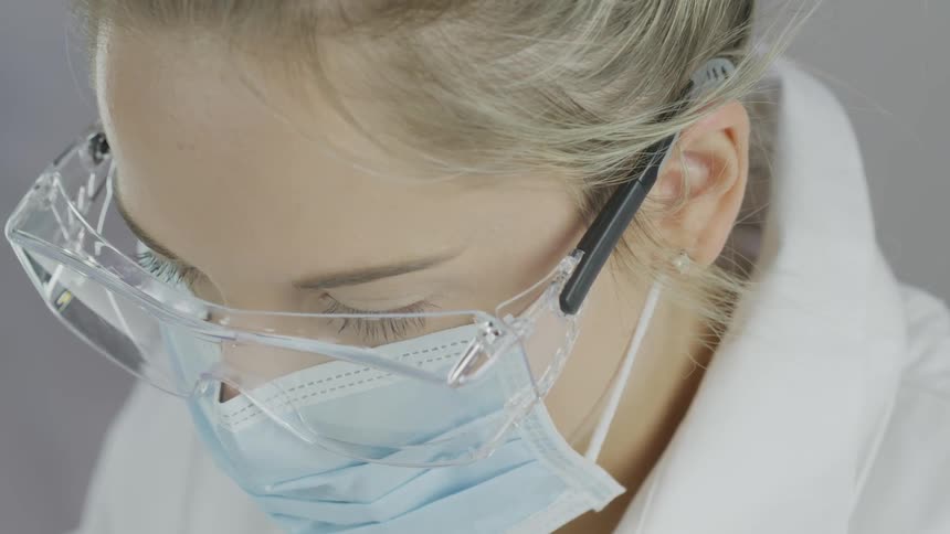 A female technician is wearing laboratory safety glasses a disposable face mask and a white lab coat as she concentrates on her work