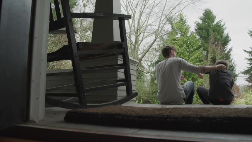 A young adult male sits with an adolescent male on the steps of a porch as the two playfully push each other and hug
