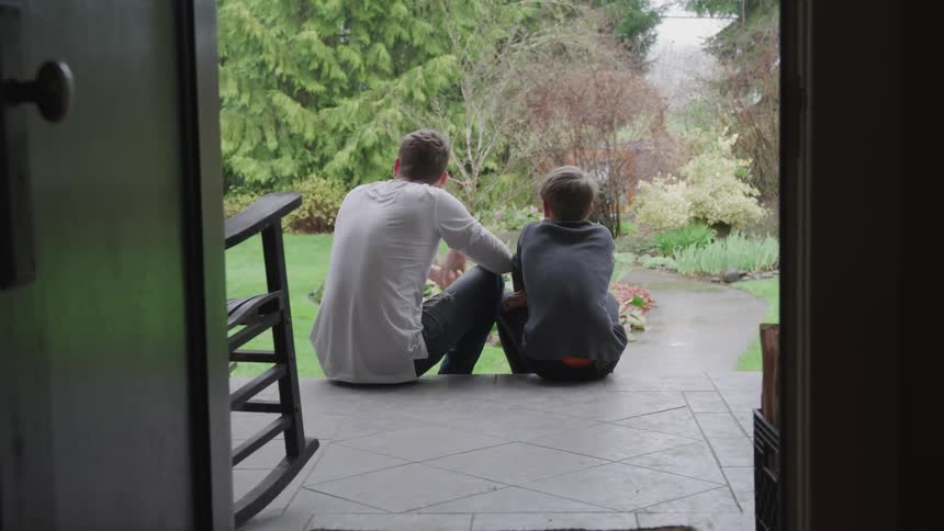 A father and son talk while sitting on the front porch of a house
