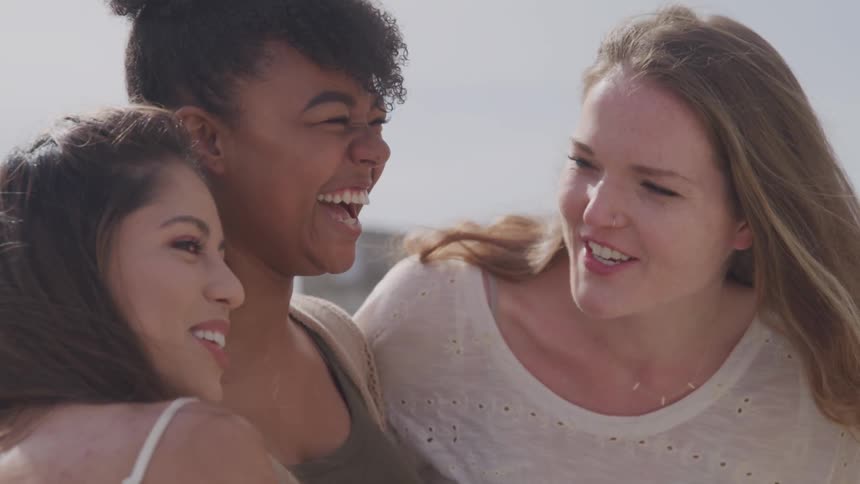Three women stand together laughing and hugging on the beach