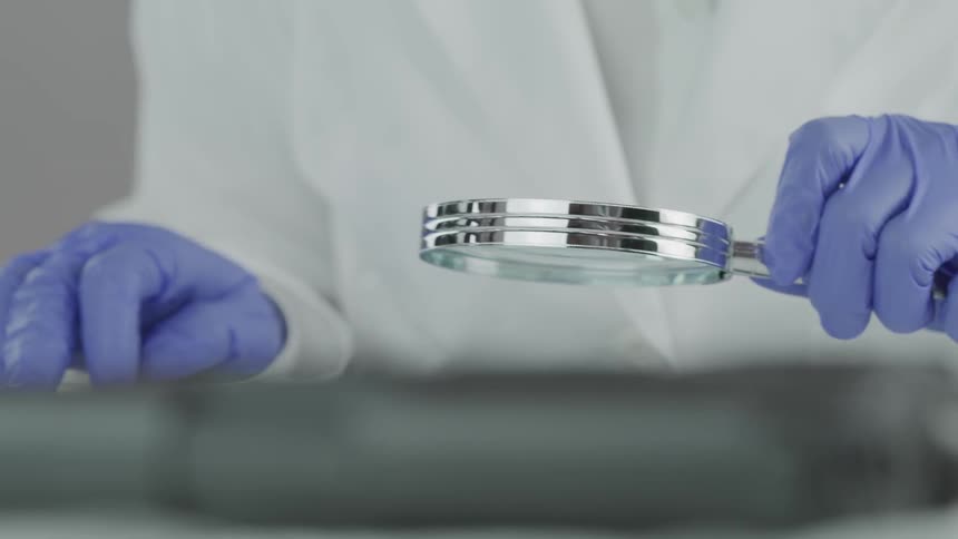 A person wearing blue disposable gloves holds a magnifying glass in their hands while inspecting an object closely