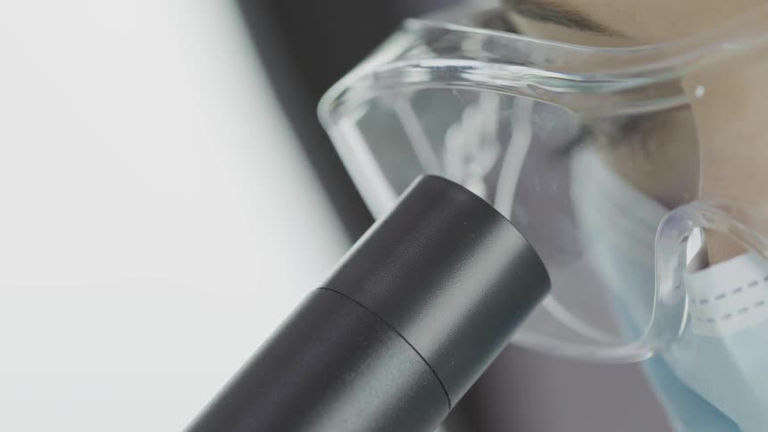 Woman wearing safety goggles and a mask looks into microscope