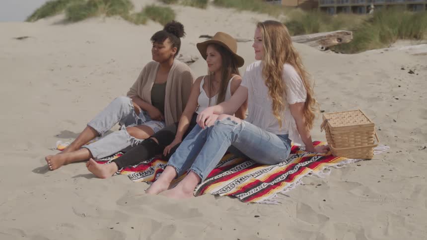 Three women sit on a blanket on a beach with a picnic basket