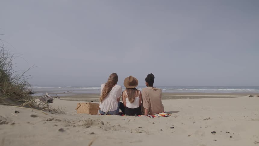 Three friends sit on the beach with a hamper