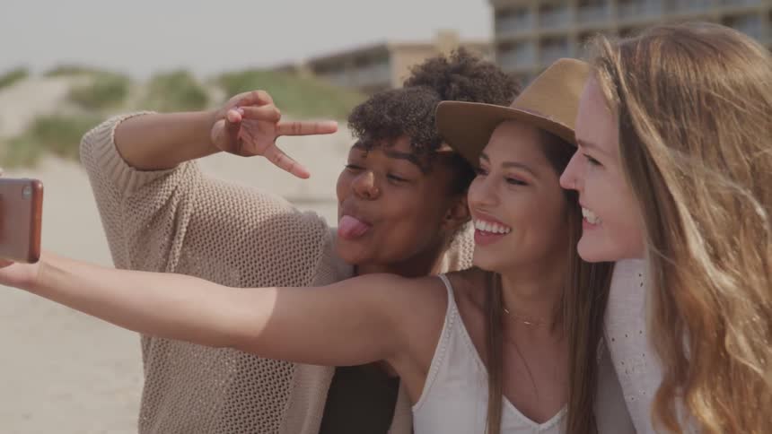 A trio of young woman lean in close to each other and smile while taking a selfie outdoors