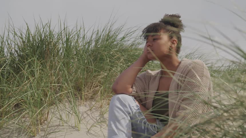 A woman sits in the grass on a beach as a breeze blows gently
