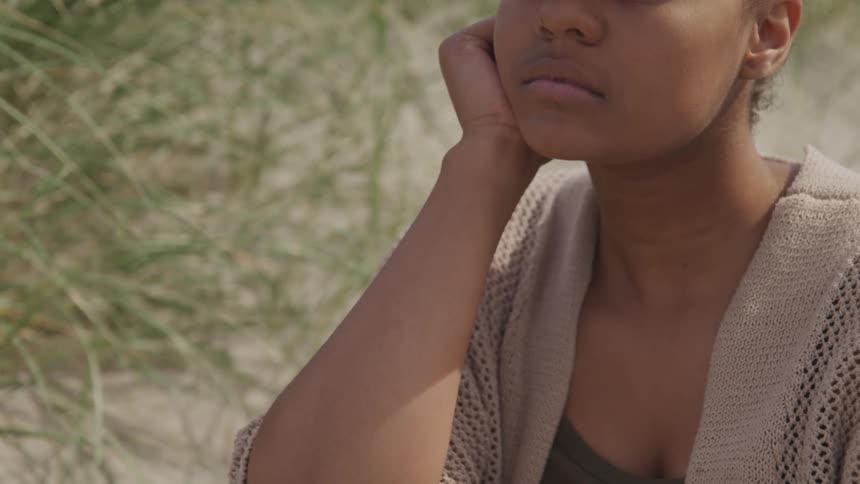 Black woman sitting on the white sand beach rests her chin on her hand during the day