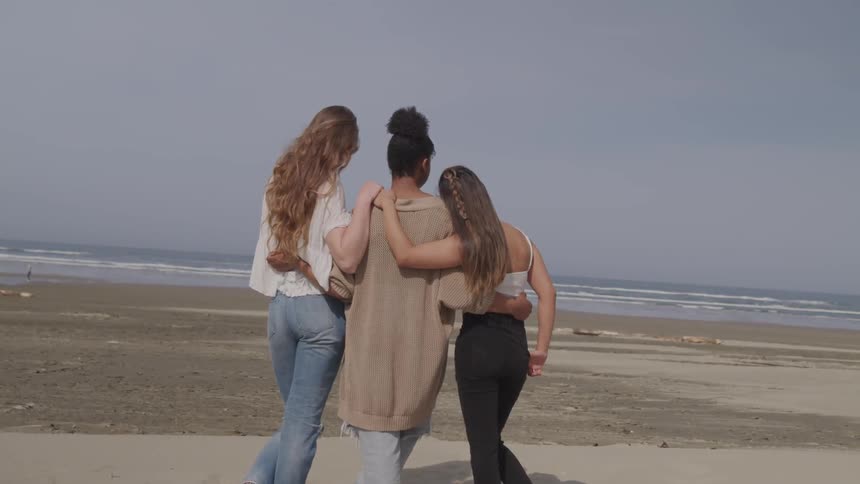 Three women stand together laughing and hugging on the beach