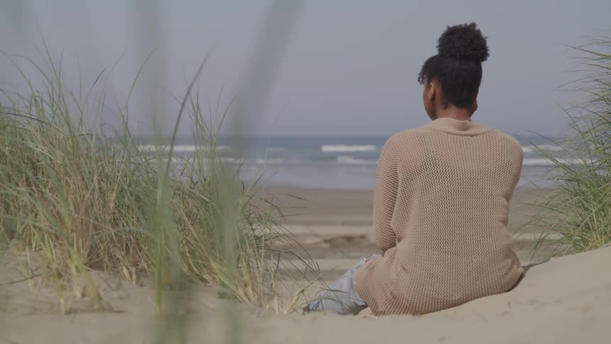 Black woman sits on the white sand beach next to grass across the ocean