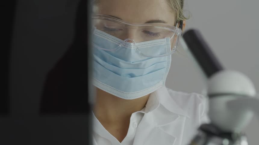 A woman with goggles and a disposable facemask looking around with a microscope in the foreground