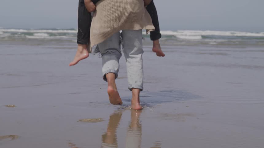 One female in a cardigan giving another female in jeans a piggyback on the beach