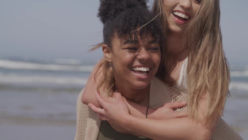 A woman giving a piggy back ride to another at the beach turns as they both laugh